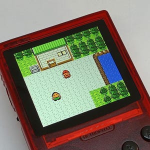 Puede incluir: Una consola de juegos port&aacute;til roja que muestra una escena de videojuego retro con gr&aacute;ficos pixelados. La pantalla muestra una vista desde arriba de un edificio, &aacute;rboles y dos personajes. El dispositivo tiene botones negros y la palabra RETROPIXEL.