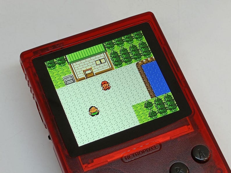 Puede incluir: Una consola de juegos port&aacute;til roja que muestra una escena de videojuego retro con gr&aacute;ficos pixelados. La pantalla muestra una vista desde arriba de un edificio, &aacute;rboles y dos personajes. El dispositivo tiene botones negros y la palabra RETROPIXEL.