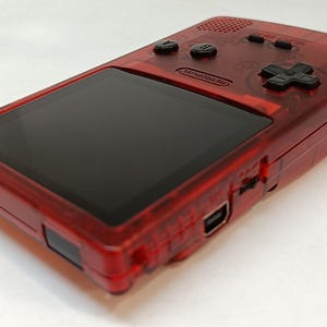 May include: A transparent red handheld gaming console with a black screen and black buttons. The console has a directional pad and two action buttons. The word "Nintendo" is visible on the device.