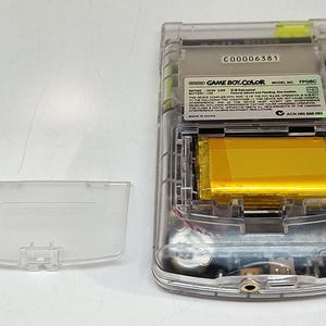Puede incluir: Una consola de juegos port&aacute;til Game Boy Color transparente con el compartimento de la bater&iacute;a abierto, revelando una bater&iacute;a dorada. La cubierta trasera de pl&aacute;stico transparente est&aacute; separada. El texto "GAME BOY COLOR" es visible.