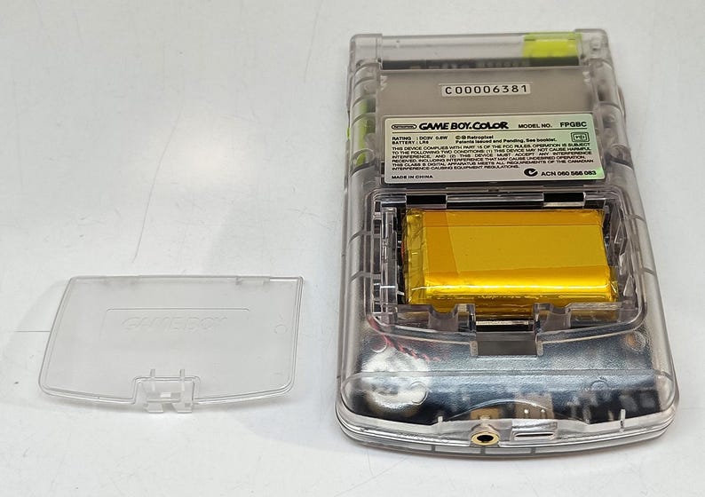 Puede incluir: Una consola de juegos port&aacute;til Game Boy Color transparente con el compartimento de la bater&iacute;a abierto, revelando una bater&iacute;a dorada. La cubierta trasera de pl&aacute;stico transparente est&aacute; separada. El texto "GAME BOY COLOR" es visible.