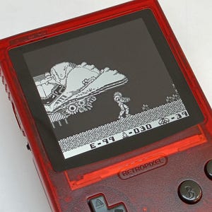 Puede incluir: Una consola de juegos port&aacute;til roja transl&uacute;cida que muestra una escena de videojuego retro en blanco y negro. La pantalla muestra un personaje pixelado y una nave espacial. El dispositivo tiene botones negros y el texto "RETROPIXEL".