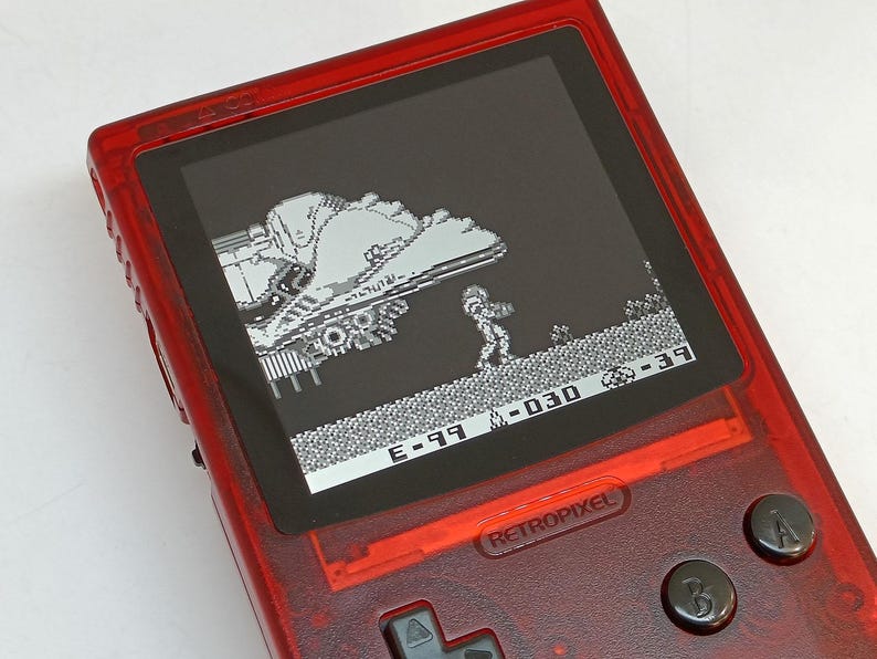 Puede incluir: Una consola de juegos port&aacute;til roja transl&uacute;cida que muestra una escena de videojuego retro en blanco y negro. La pantalla muestra un personaje pixelado y una nave espacial. El dispositivo tiene botones negros y el texto "RETROPIXEL".