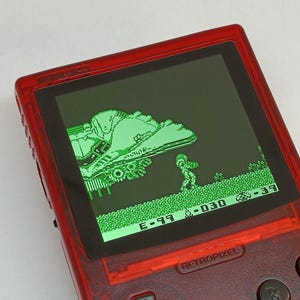 Puede incluir: Una consola de juegos port&aacute;til Retropixel roja que muestra una escena de videojuego te&ntilde;ida de verde. El juego presenta un personaje pixelado y un veh&iacute;culo en un paisaje. El dispositivo tiene botones negros y una pantalla con detalles del juego.