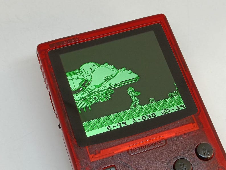 Puede incluir: Una consola de juegos port&aacute;til Retropixel roja que muestra una escena de videojuego te&ntilde;ida de verde. El juego presenta un personaje pixelado y un veh&iacute;culo en un paisaje. El dispositivo tiene botones negros y una pantalla con detalles del juego.