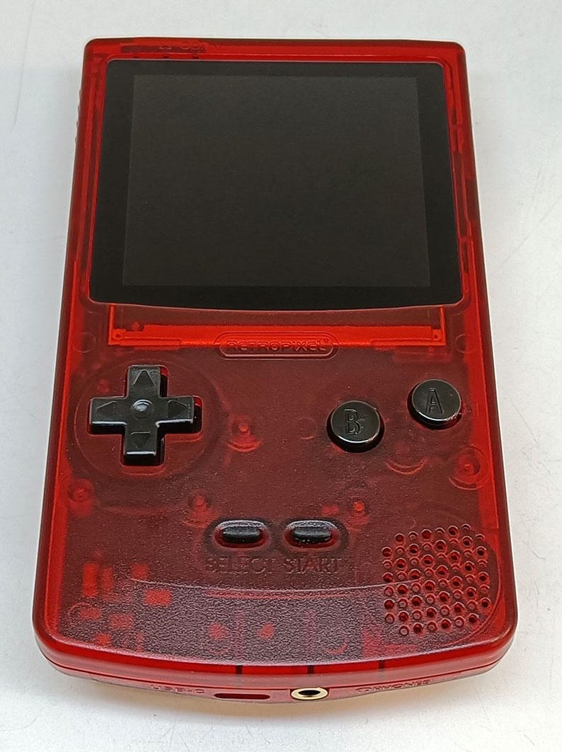 May include: A transparent red handheld gaming console with a black screen and black buttons. The console has a directional pad, two action buttons, and select/start buttons. The text "RETROPIXEL" is visible on the device.