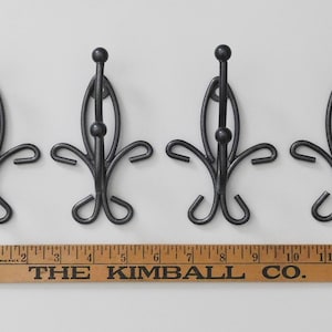 May include: Four black metal coat hooks with a decorative design. Each hook has a rounded top and a curved base. A wooden ruler with the text "THE KIMBALL CO." is below the hooks. The hooks are approximately 5 inches in height.