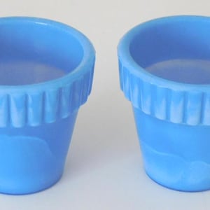 May include: Two matching light blue glass vessels with a tapered, cylindrical shape. Each vessel features a wide, fluted rim with vertical ridges encircling the top edge. The glass has a semi-translucent, smooth finish with subtle variations in color intensity.