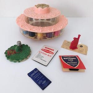 May include: A pink plastic rotating sewing thread organizer with multiple spools, a green crocheted pincushion with a metal thimble, a Wonder Potato Chips stitch kit, a red plastic tool, a National Life and Accident Insurance Co. card, and a blue Guardian needle packet.