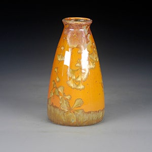 May include: A ceramic vase with a tapered neck and a glossy orange glaze. The surface features crystalline patterns in shades of gold and brown that resemble floral or leaf-like formations, with a darker, textured base in earthy brown tones.