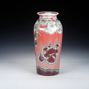 May include: A red ceramic vase with a floral design. The vase has a white glaze with green and brown accents. The flowers are stylized and have a whimsical feel.