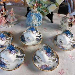 Può includere: Un set di tazze e piattini in porcellana con un motivo floreale blu e viola. Le tazze sono disposte su una tovaglia bianca, con statuine decorative sullo sfondo. Le tazze hanno bordi dorati.