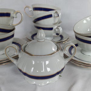 May include: A white porcelain tea set featuring a sugar bowl with a lid and multiple teacups with matching saucers. Each piece is decorated with a bold navy blue horizontal band and thin gold trim along the edges and handles.