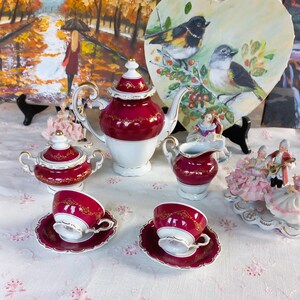 May include: A porcelain tea set featuring a teapot, a lidded sugar bowl, a creamer, and two teacups with saucers. Each piece is finished in deep burgundy and white with intricate gold filigree detailing along the rims and central bands.