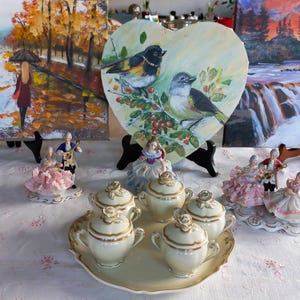 Puede incluir: Una colección de objetos decorativos, que incluye una pintura en forma de corazón de pájaros, figuritas de porcelana y un juego de té con una bandeja. El juego de té es de color crema con ribetes dorados.