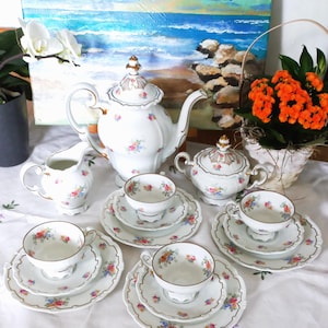 May include: A white porcelain tea set featuring a teapot, a lidded sugar bowl, a creamer, and four teacups with matching saucers and plates. Each piece is decorated with delicate multi-colored floral patterns and gold-trimmed scalloped edges.