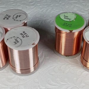 May include: Five spools of copper-colored craft wire, each labeled Bead Landing with 24 yd and 21.9 m printed on the top. The spools are cylindrical with a metallic finish, arranged in a group. The labels are white or light green in color.