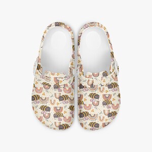 May include: A pair of cream-colored clogs with a repeating pattern of bees, rainbows, and floral accents. The clogs have a white interior and adjustable straps. These shoes are designed for comfort and style.