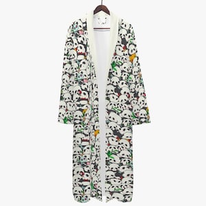 Puede incluir: Bata larga y abierta por delante con un estampado repetido de caras de panda en blanco y negro. La bata tiene un interior y un cuello blancos, y está colgada de una percha de madera. Las caras de los pandas tienen varias expresiones y están intercaladas con bambú verde.