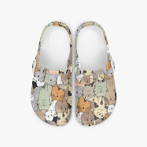 May include: A pair of clogs with a repeating pattern of cartoon cats in various colors, including gray, orange, and brown. The clogs have white soles and adjustable straps. The overall design is playful and whimsical.