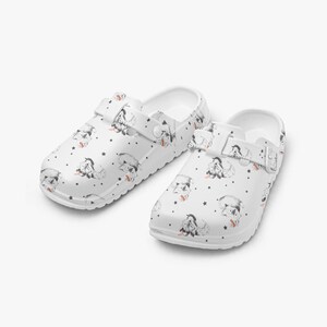 May include: White clogs with a repeating pattern of cartoon dogs and black stars. The clogs have adjustable straps and are made of a durable material. The dog illustrations have red accents.