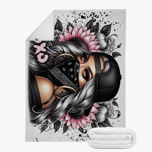 May include: White blanket with a detailed illustration of a woman with long hair, wearing a bandana and a baseball cap. The design includes pink flowers and the text "OX" in pink. The blanket is folded at the bottom.