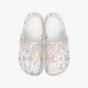 May include: White clogs featuring a repeating pattern of cartoon cats in pink, yellow, and grey. The clogs have a light beige strap and a white interior. The clogs are suitable for indoor and outdoor use.