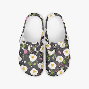May include: A pair of black clogs with a repeating pattern of fried eggs, herbs, and vegetables. The clogs have white soles and straps. The egg yolks are yellow, and the vegetables are red, pink, and green.