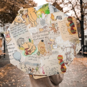 May include: An octagonal umbrella featuring a collage of Winnie the Pooh characters and quotes. The design includes illustrations of Pooh, Piglet, Tigger, and Eeyore, with text overlays and honey pots. The umbrella is beige and brown.
