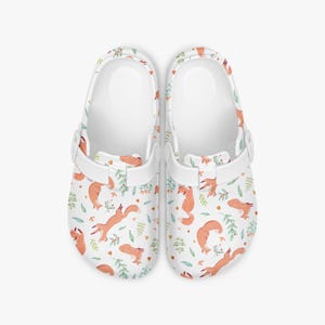 May include: A pair of white clogs with a repeating pattern of orange foxes, green leaves, and small orange hearts. The clogs have white straps and a white interior.