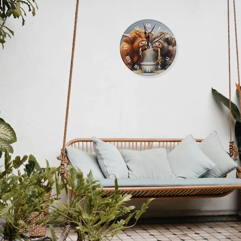 May include: A clock featuring two Highland cows eating ramen noodles from a bowl. The clock has a white background with black Roman numeral hour markers and black hands. The cows are brown and white with black noses and hooves. The clock is hanging on a white wall above a light brown wicker swing bench with light blue cushions.