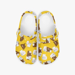 May include: A pair of yellow clogs with a repeating pattern of cartoon poop and toilet paper rolls. The clogs have a white interior and a brown strap.
