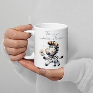 May include: White ceramic mug featuring a cartoon zebra holding a daisy. The zebra has black and white stripes and a brown mane. The mug has the text "Für einen wundervollen Menschen" in a script font.