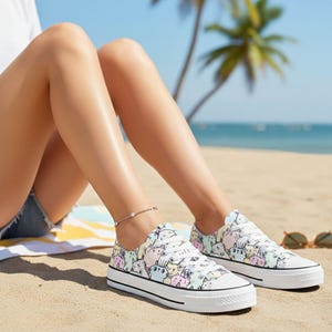 Può includere: Scarpe da ginnastica in tela bianca con un motivo a gatti colorati. Le scarpe basse hanno lacci bianchi e una striscia nera sulla suola. Le scarpe sono indossate su una spiaggia sabbiosa. Un accessorio divertente e giocoso.