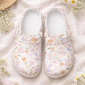 Puede incluir: Un par de zuecos blancos con un estampado floral en rosa, melocotón y azul. Los zuecos tienen un interior blanco y correas ajustables. El diseño floral cubre todo el zapato, creando un aspecto decorativo.