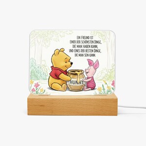 May include: A decorative night light featuring Winnie the Pooh and Piglet, with the word "HUNNY" on a honey pot. The light has a wooden base and a printed image with the text "EIN FREUND IST...DIE MAN SEIN KANN."