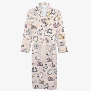 May include: A light beige bathrobe featuring a repeating pattern of cartoon cats in various poses and colors, along with small fish and the word "meow". The robe has long sleeves, a belt, and a shawl collar.