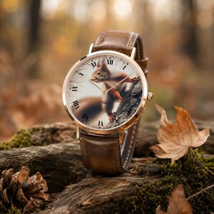 May include: A wristwatch with a brown leather strap and a rose gold-toned metal case. The watch face features a detailed illustration of a squirrel on a tree branch, black Roman numeral hour markers, and thin gold-toned hands with the text Nihis.