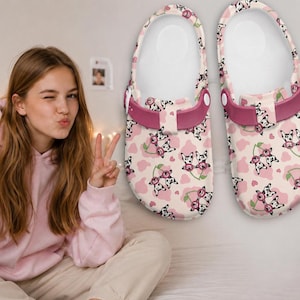 May include: A pair of pink and white clogs with a Dalmatian print. The clogs have a pink strap and a white interior. The Dalmatian print includes pink hearts and the clogs are on a white background.