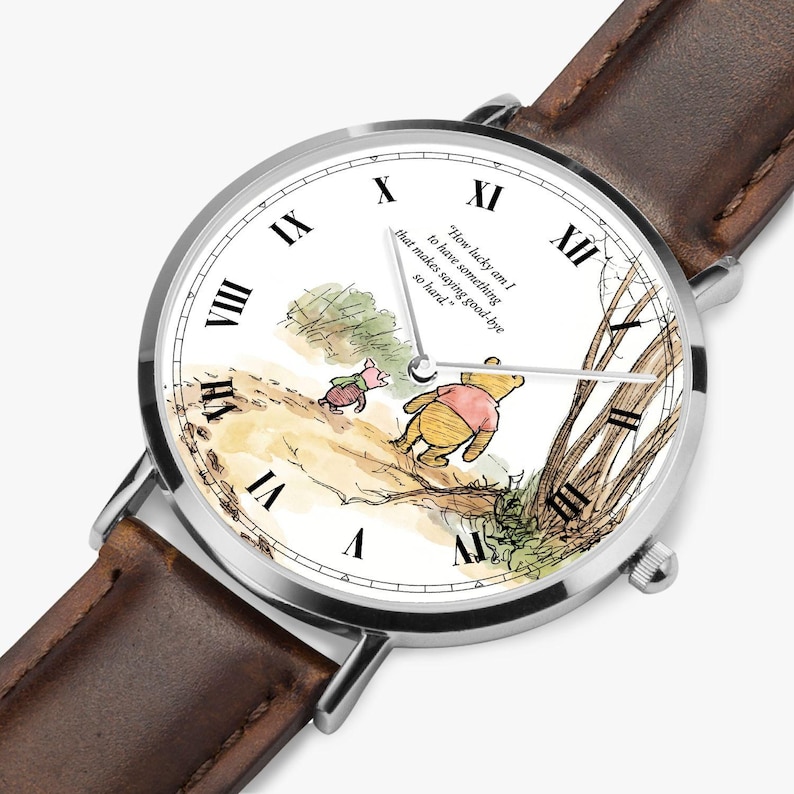 Pode incluir: Um rel&oacute;gio de pulso prateado com uma bracelete de couro castanho. O mostrador do rel&oacute;gio apresenta uma ilustra&ccedil;&atilde;o em aquarela de Winnie the Pooh e Piglet, com a cita&ccedil;&atilde;o "How lucky am I to have something that makes saying good-bye so hard."