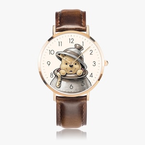 Puede incluir: Un reloj de pulsera dorado con una correa de cuero marrón. La esfera del reloj presenta una ilustración de dibujos animados de Winnie the Pooh en un tarro de miel. El reloj tiene números y agujas negros.