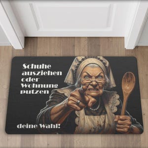May include: Black rectangular doormat featuring a stern-looking elderly woman with a wooden spoon, pointing. Text reads "Schuhe ausziehen oder Wohnung putzen deine Wahl!" in white. The woman wears a bonnet and apron.