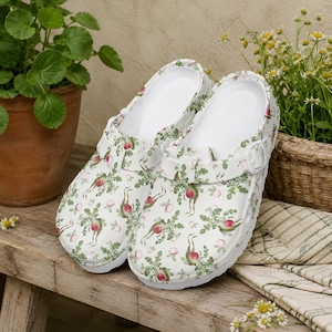 May include: A pair of white clogs with a radish pattern. The clogs have a white interior and a strap with a buckle. The radish design includes red radishes with green leaves and stems, scattered across the surface.