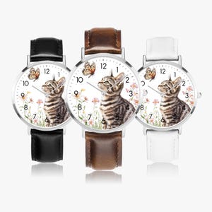 May include: Three wristwatches with silver cases and leather straps in black, brown, and white. Each watch face features a tabby cat illustration with a butterfly and floral accents. The watches display the numbers 1 to 12.