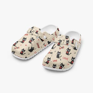 Puede incluir: Zuecos beige con suela blanca, con un estampado repetido de gatos negros, notas musicales y corazones. La palabra "LOVE" está impresa en el lateral. Los zuecos tienen correas ajustables.