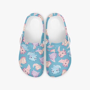 May include: A pair of blue clogs featuring a repeating pattern of cartoon axolotls in pink, purple, and blue. The clogs have white inner soles and adjustable straps. The axolotls have smiling faces and are in various poses.