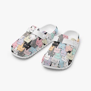 May include: A pair of white clogs with a colorful pattern of cat faces. The clogs have a white strap and a white sole. The cat faces are in various colors, including pink, black, yellow, and gray.