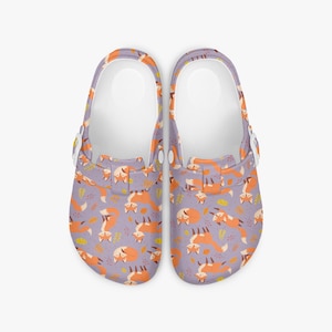 May include: A pair of clogs with a repeating pattern of orange foxes and yellow leaves on a lavender background. The clogs have white soles and adjustable straps. The clogs are designed for comfort and style.