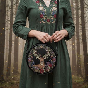 May include: A round black crossbody bag featuring a detailed illustration of a deer with large antlers, surrounded by colorful flowers and foliage. The bag has a black strap and is held against a green dress with floral embroidery.