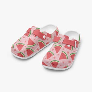 Könnte beinhalten: Paar weiße Clogs mit einem rosa Wassermuster. Die Clogs haben einen rosa Riemen und eine weiße Sohle. Die Wassermelonenstücke sind rot mit grünen Schalen und schwarzen Samen. Der Hintergrund ist hellrosa mit weißen Punkten.